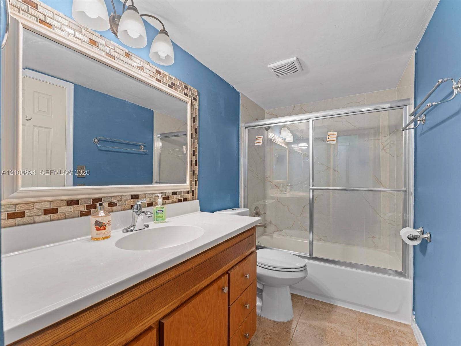 Bathroom featuring a wood-finish vanity with a white countertop, a framed wall mirror with mosaic tile trim, and a bathtub with sliding glass doors and surround tile