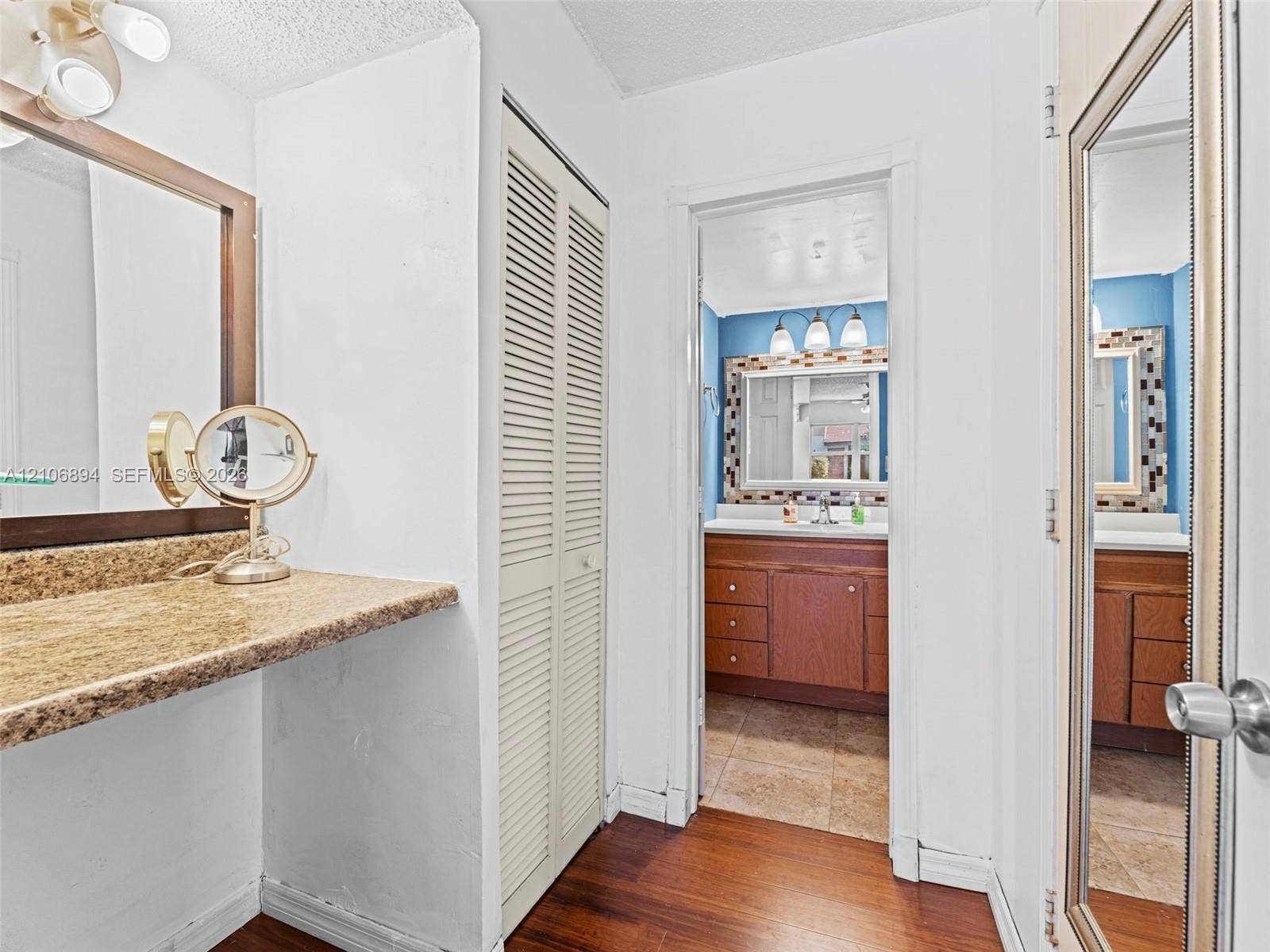 Dressing area featuring stone-finish countertop, wood-finish flooring, and floor-to-ceiling louvered closet doors, leading to a vanity area equipped with wood cabinetry and tile flooring