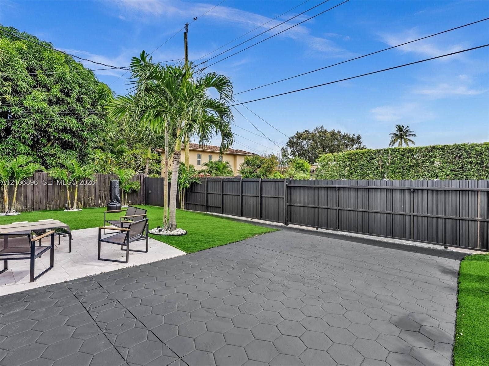 Expansive backyard featuring a concrete patio with a hexagonal pattern, synthetic turf lawn, perimeter wood privacy fencing, and mature palm trees