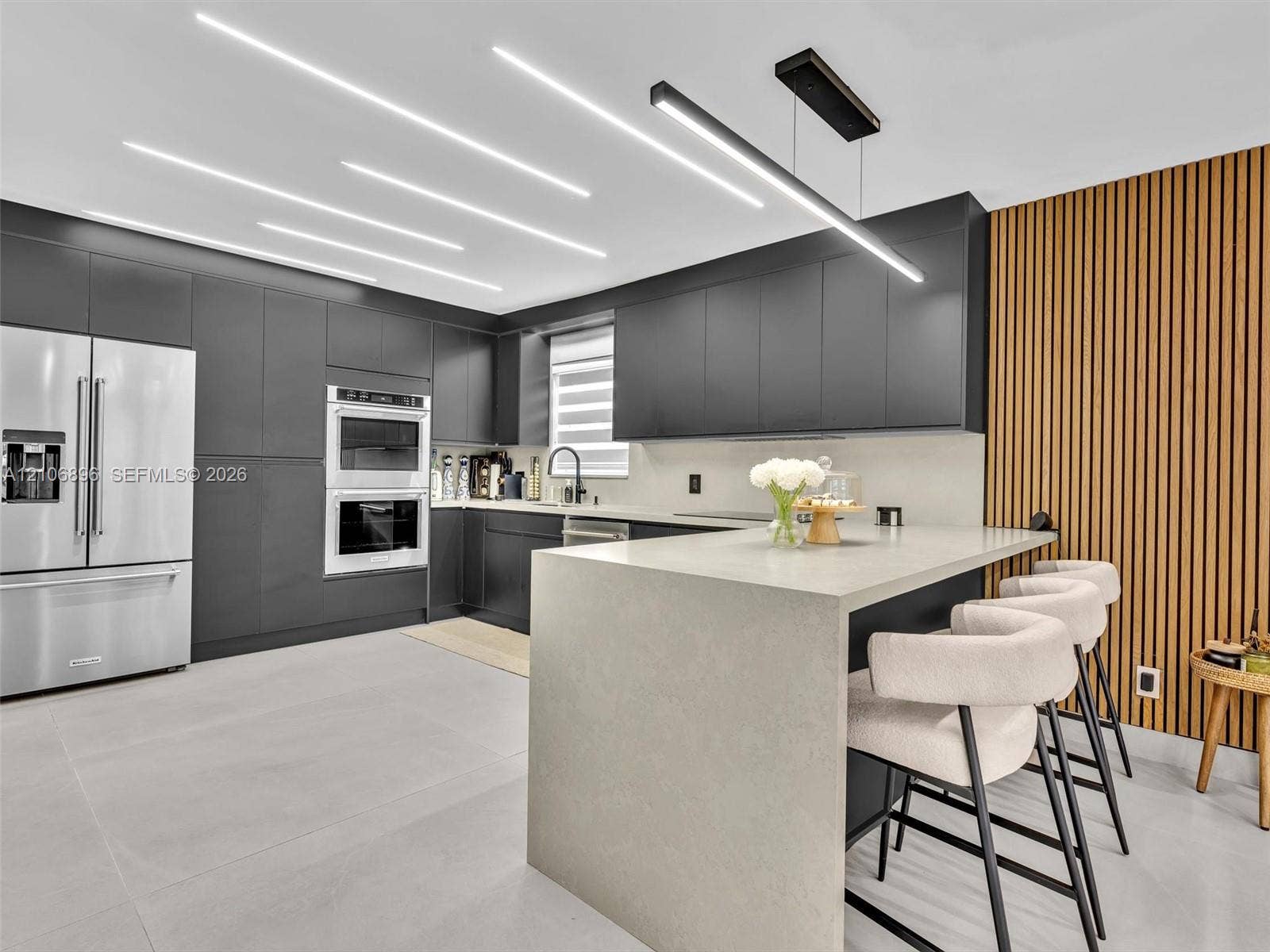 Modern kitchen featuring flat-panel cabinetry, a waterfall-edge stone island, linear LED ceiling lighting, integrated double wall ovens, and a vertical wood-slat accent wall