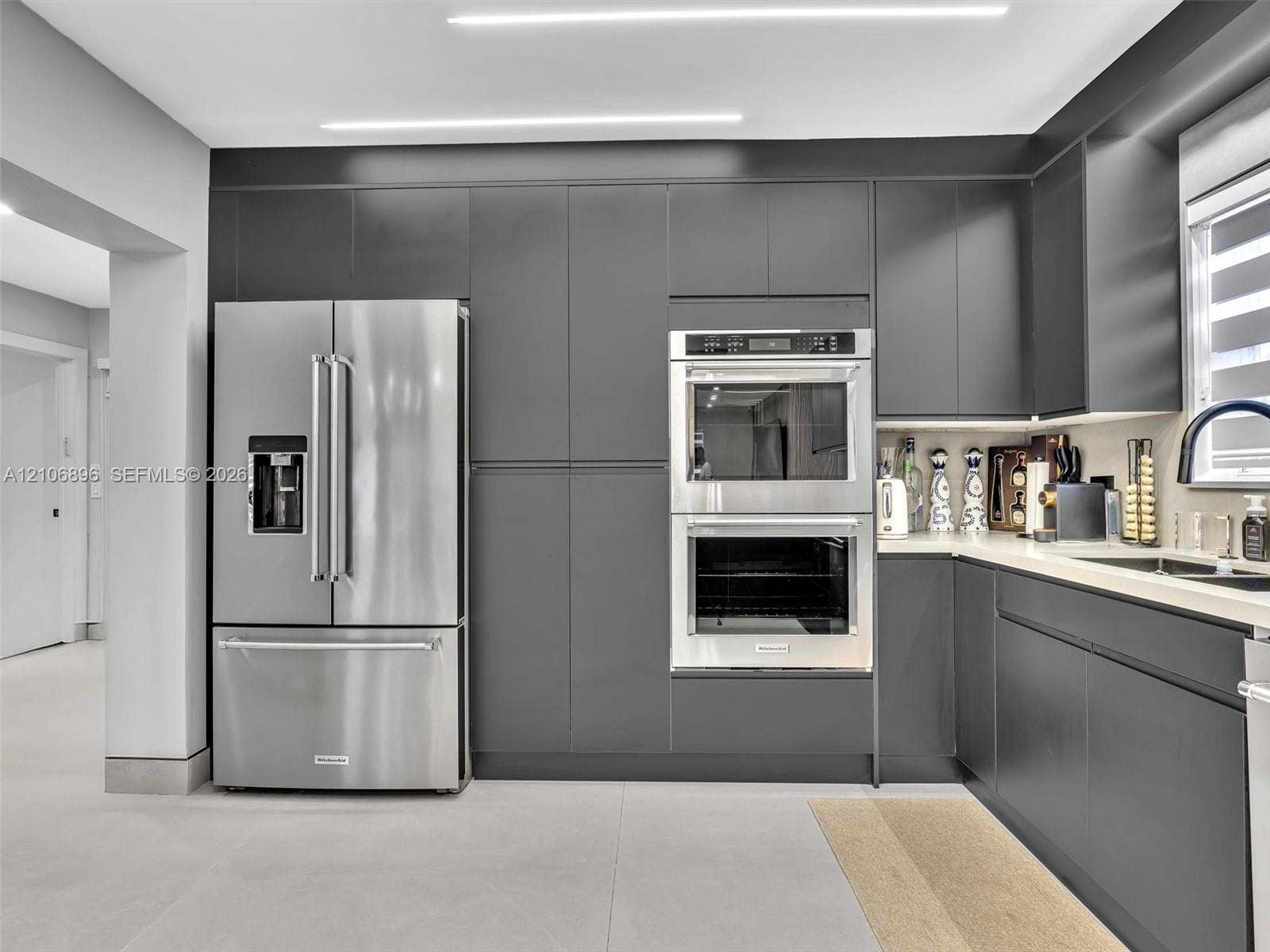 Sleek charcoal cabinetry, stainless steel double wall ovens, French door refrigerator, light-toned countertop, and integrated linear overhead lighting