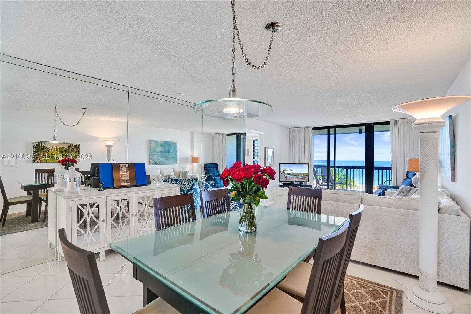 510 N Ocean Boulevard, #308, Pompano Beach, FL 33062 Open-concept main living area featuring large-format ceramic tile flooring, a wall-to-wall mirrored accent wall, suspended glass-shade pendant lighting, and a floor-to-ceiling glass slider with balcony access