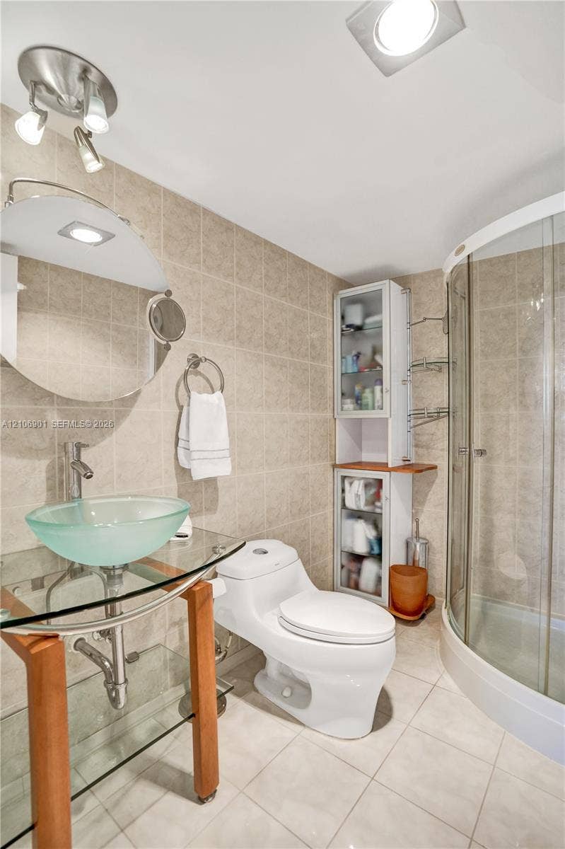 510 N Ocean Boulevard, #308, Pompano Beach, FL 33062 Glass vessel sink with chrome hardware, wood-frame pedestal vanity, curved glass-enclosed shower, built-in cabinetry, and ceramic tile wall and floor surfaces
