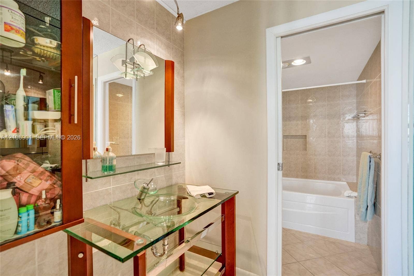 510 N Ocean Boulevard, #308, Pompano Beach, FL 33062 Vanity featuring a glass countertop, integrated vessel sink, and wood-finish frame, complemented by a wall-mounted mirror, glass shelving, and tile shower surround with a built-in recessed niche