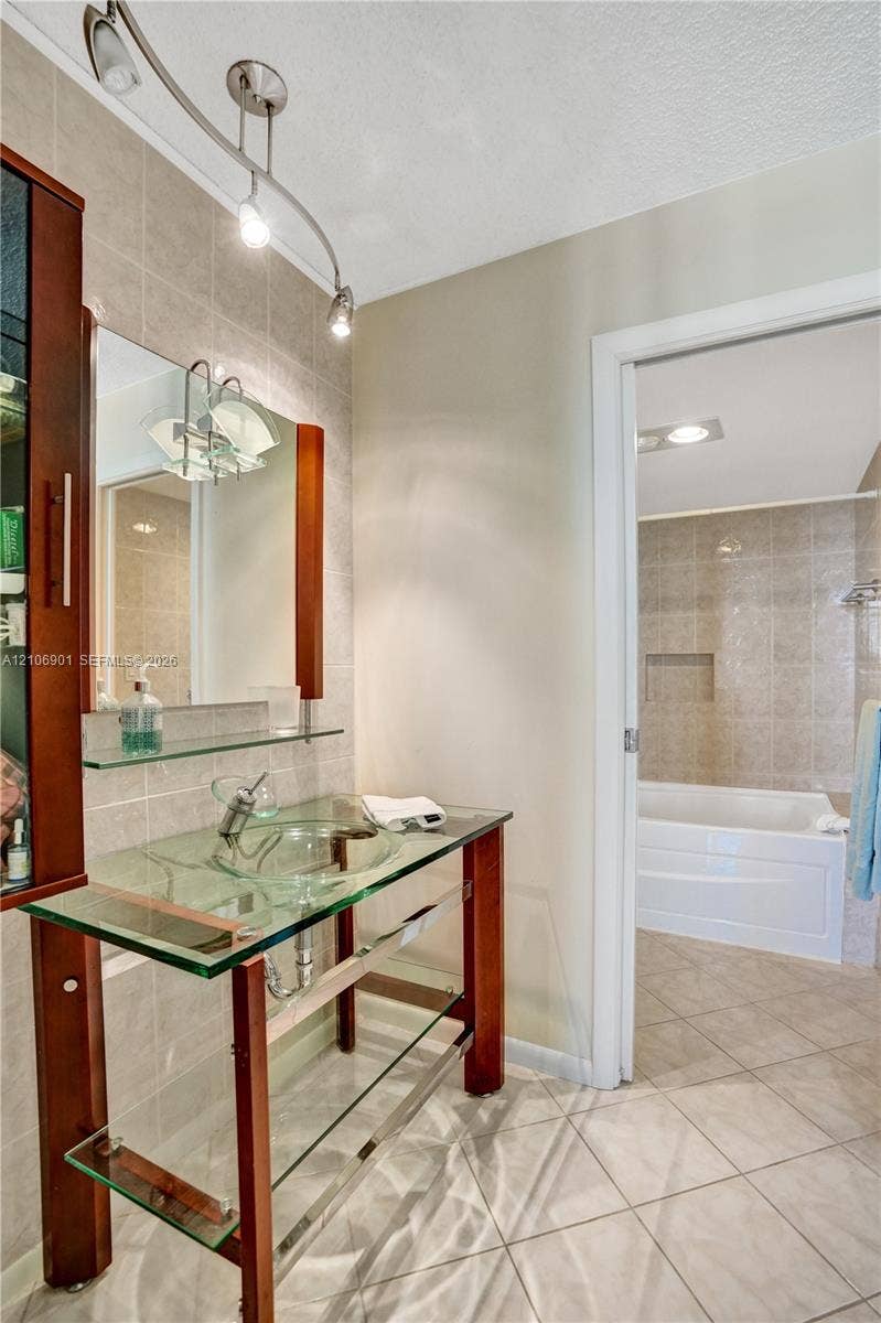 510 N Ocean Boulevard, #308, Pompano Beach, FL 33062 Glass vessel sink vanity featuring a wood-finish frame, clear glass shelving, wall-mounted track lighting, and a ceramic-tiled tub enclosure