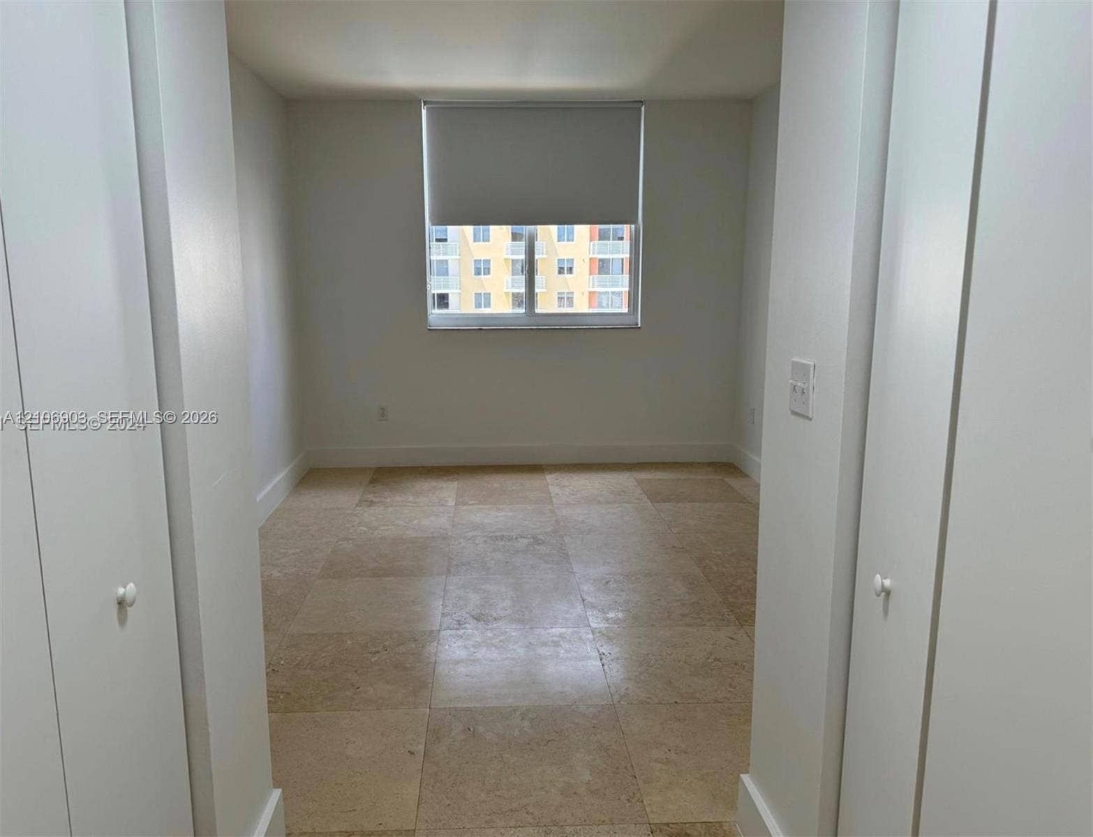 Neutral-toned tile flooring, dual closets with white swing doors, and a central window with a roller shade