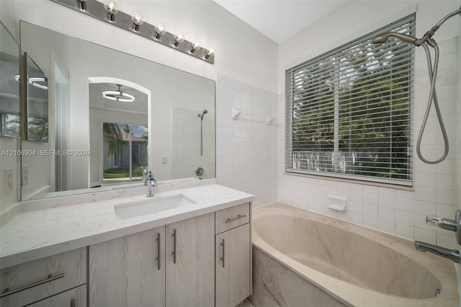 297 Conservation Drive, Weston, FL 33327 Bathroom featuring a light-toned wood-finish vanity with a rectangular sink, quartz countertop, large wall-to-wall mirror, oval soaking tub, and subway tile wall surround