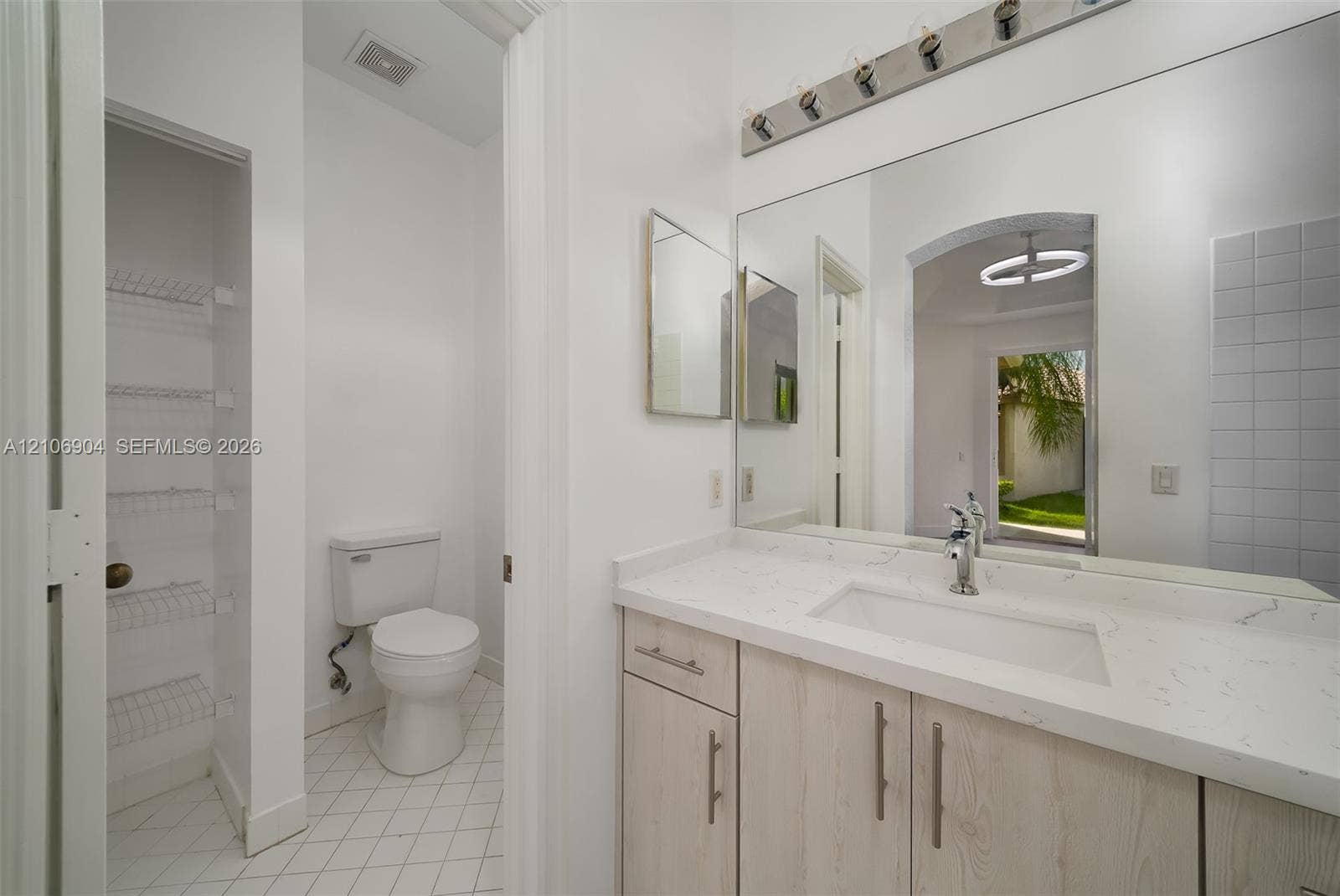 297 Conservation Drive, Weston, FL 33327 Vanity featuring a light wood-finish cabinet, quartz countertop, and integrated sink, complemented by a large wall-mounted mirror, multi-bulb lighting fixture, and built-in shelving