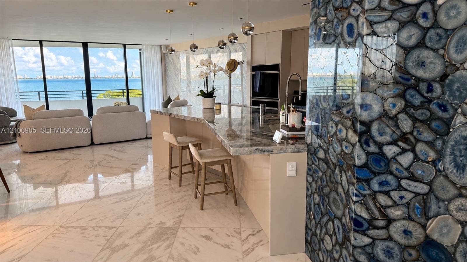 720 NE 69th Street, #10N, Miami, FL 33138 Polished stone flooring, expansive floor-to-ceiling windows with water scenery, custom waterfall-edge kitchen island, decorative blue agate stone accent wall, and built-in stainless steel appliances