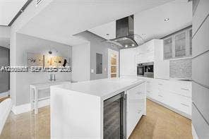 Modern kitchen featuring a central island with an integrated wine cooler, sleek white cabinetry, stainless steel range hood, built-in wall oven, and light-tone flooring