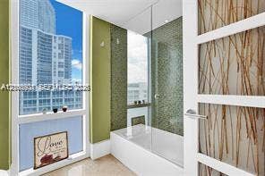 Bathroom featuring a glass-enclosed soaking tub, decorative mosaic tile surround, floor-to-ceiling windows, and a multi-panel door with integrated botanical-style accents