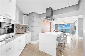 Modern kitchen featuring white flat-panel cabinetry, a central island with a solid-surface countertop, stainless steel vent hood, built-in coffee system, and polished tile flooring