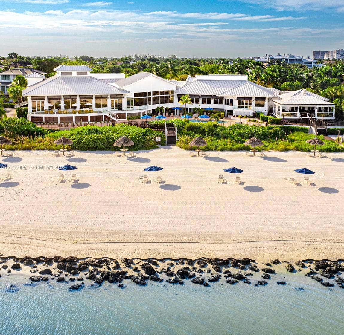331 Wild Orchid Lane, Marco Island, Marco Island, FL 34145 Direct beach access featuring a white sand shoreline, a structural stone seawall, a sprawling coastal estate with a metal roof, and manicured landscaping