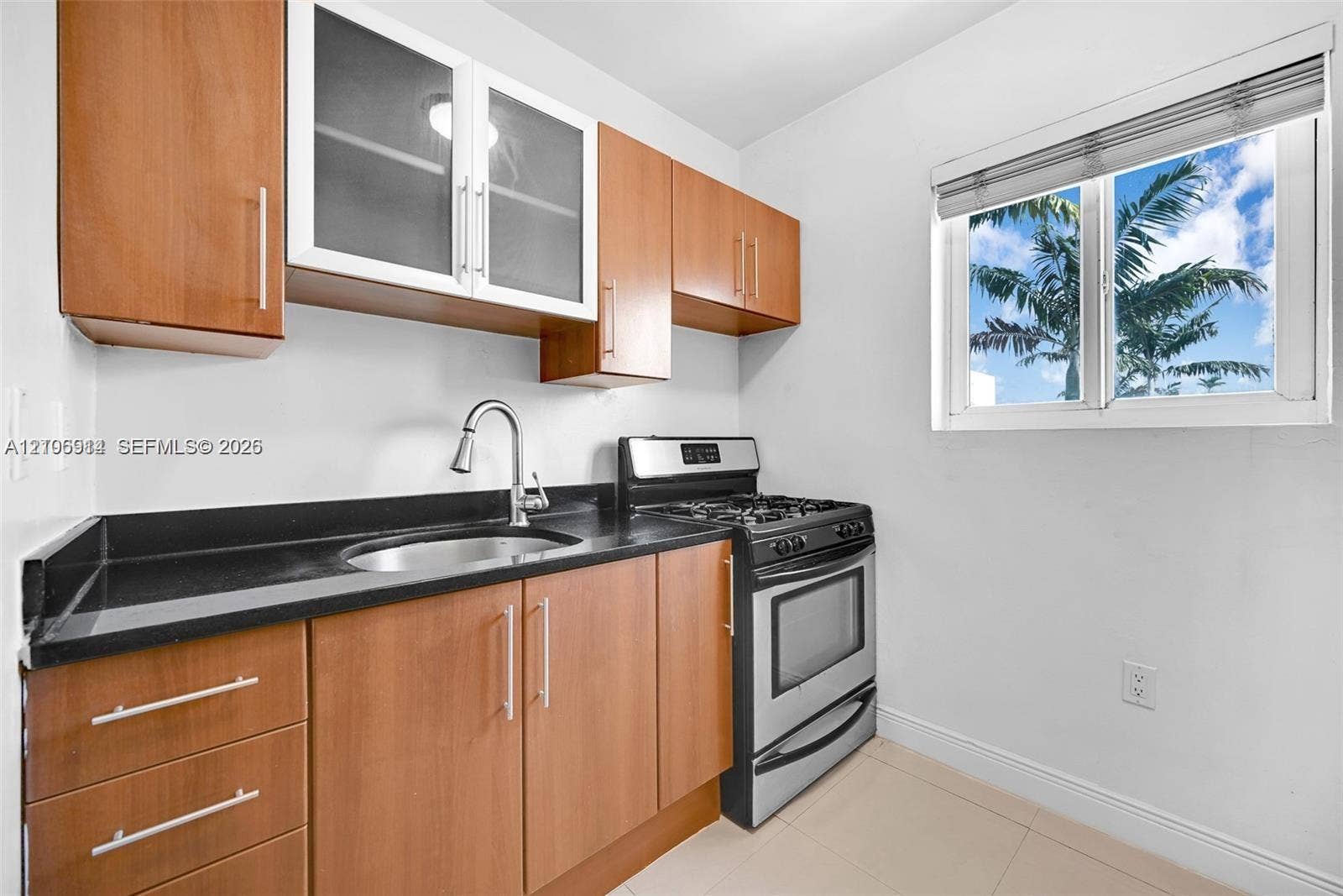320 85th Street, #20, Miami Beach, Miami Beach, FL 33141 Kitchen featuring wood-finish cabinetry with glass-front accents, dark countertops, integrated stainless steel gas range, and a single-basin undermount sink with a pull-down faucet