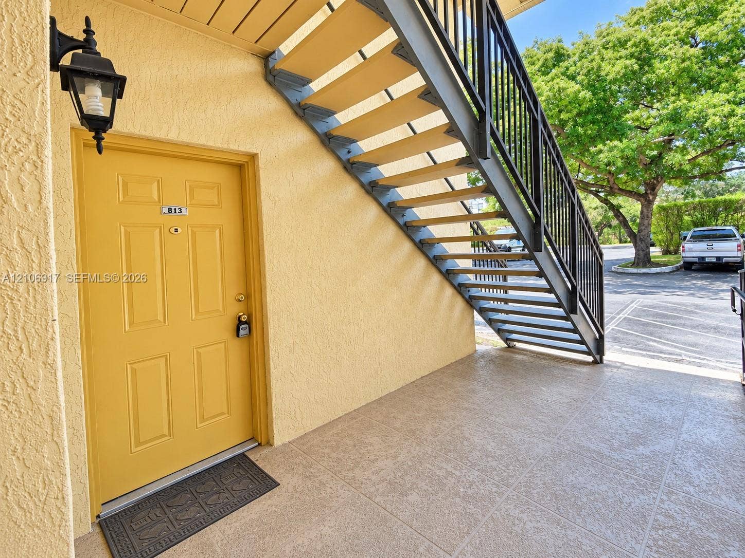 Exterior entry featuring a textured-finish door, attached decorative wall lantern, tile-style hardscaping, and black metal staircase