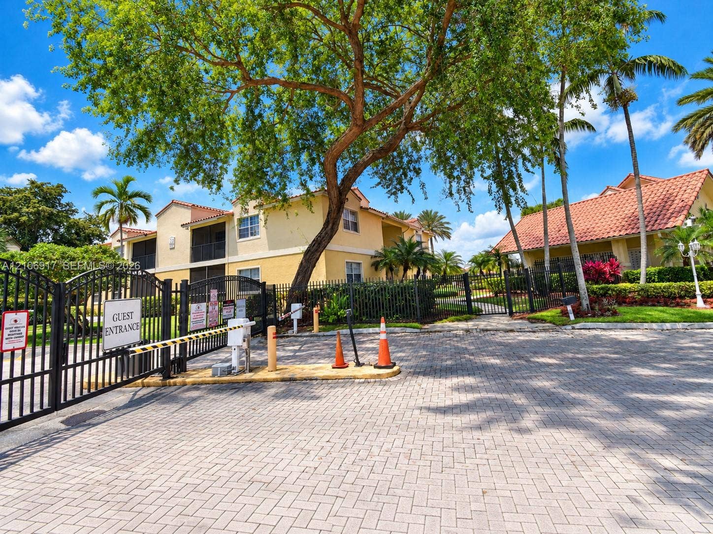 Gated community entrance featuring a paver-stone driveway, automated security gate, and mature landscaping