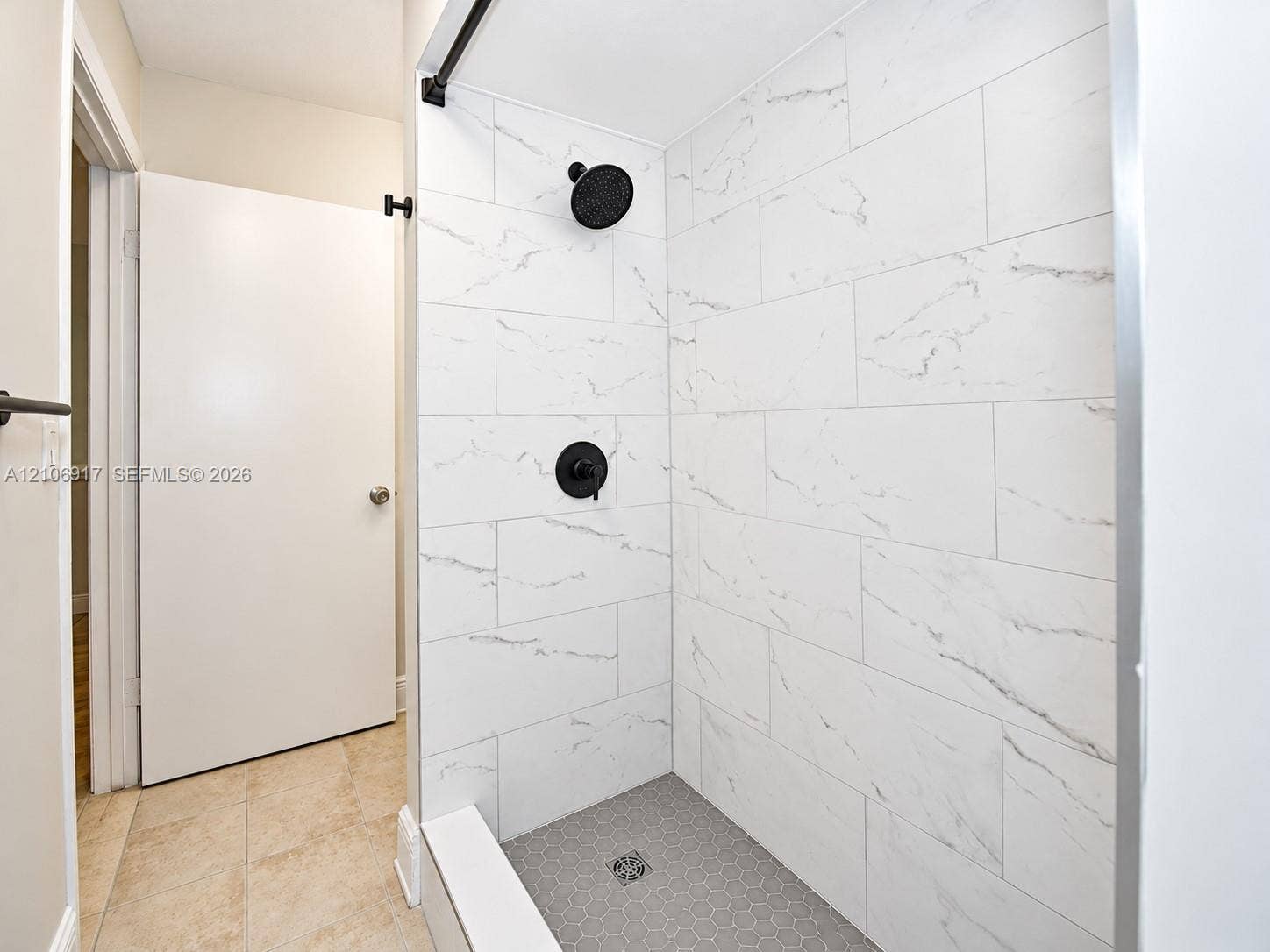 Walk-in shower featuring large-format white stone-finish wall tile, grey hexagonal floor tile, matte black wall-mounted shower head, and matte black single-handle pressure balance valve
