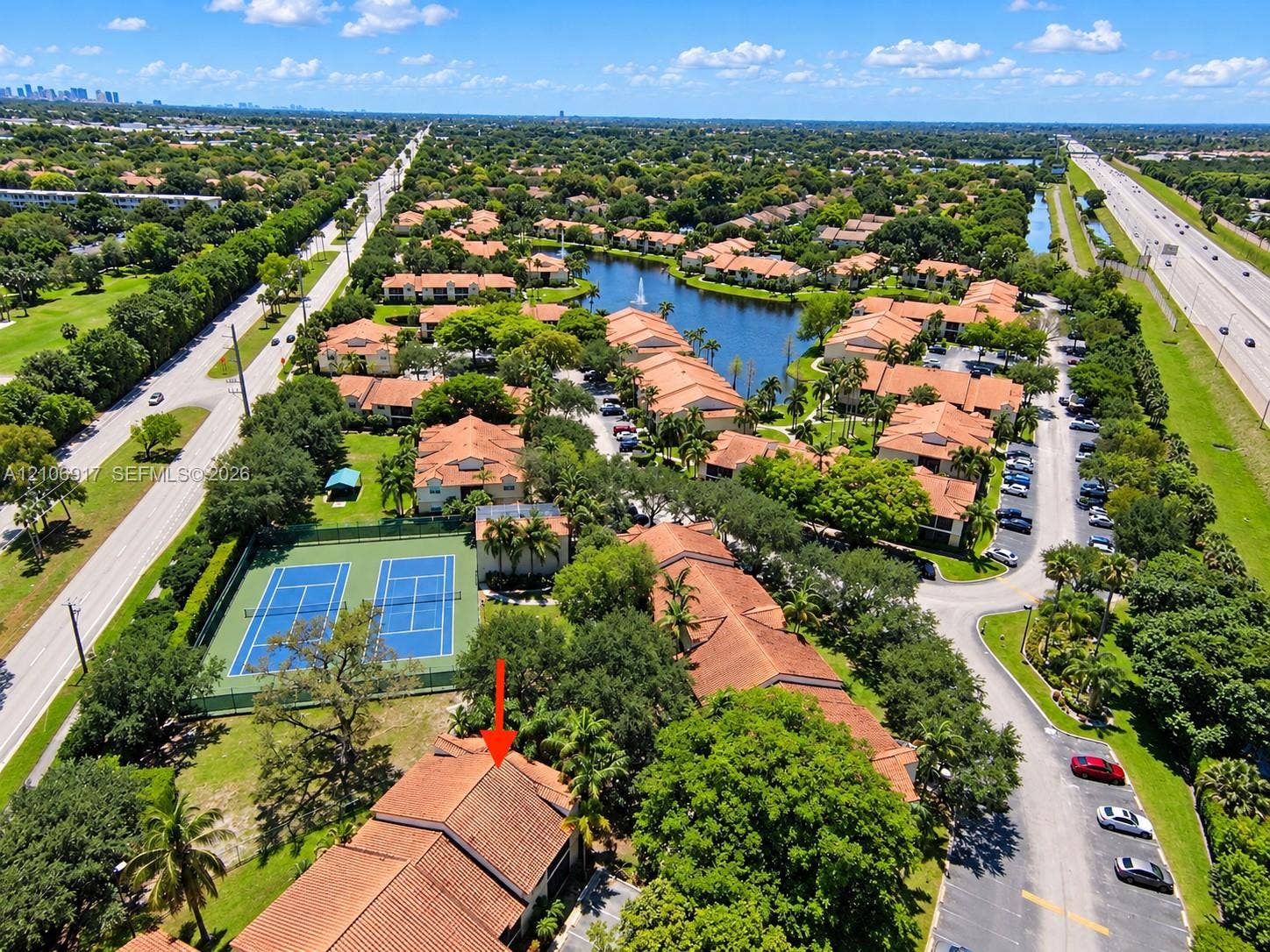 Aerial perspective of residential complex featuring tile roofing, tennis courts, a central lake with fountain, and mature landscaping