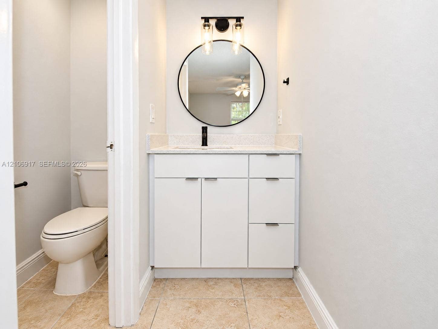 White vanity featuring a light-toned solid surface countertop, matte black faucet, circular mirror, and multi-light wall fixture with tile flooring