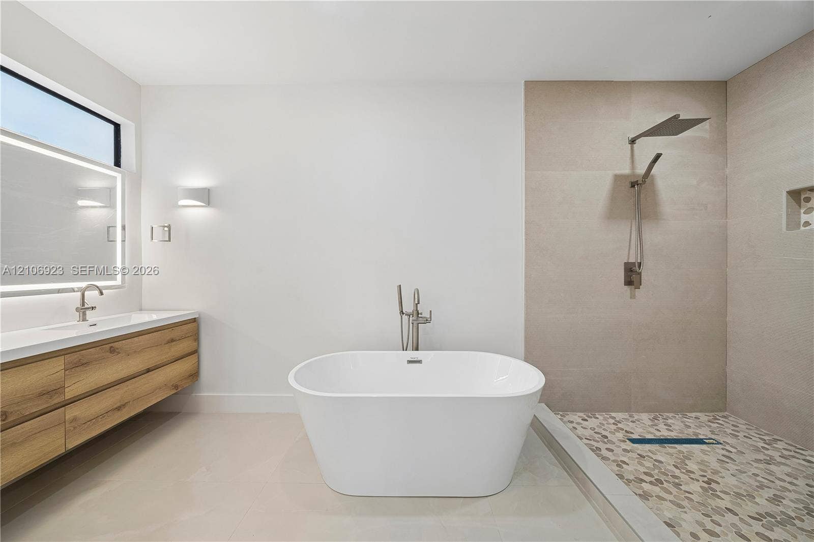 870 Superior Street, Opa Locka, FL 33054 Freestanding soaking tub with floor-mounted faucet, floating wood-finish vanity, backlit mirror, walk-in shower with pebble-stone flooring, and wall-mounted rainfall showerhead