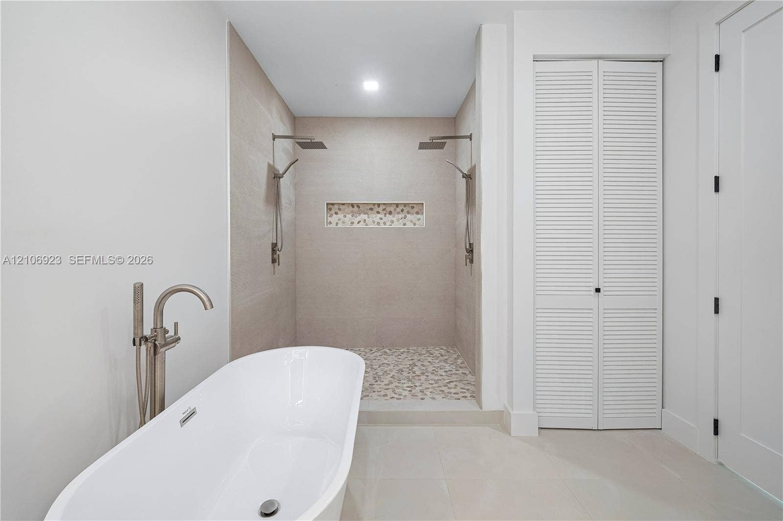 870 Superior Street, Opa Locka, FL 33054 Freestanding soaking tub with metallic floor-mounted faucet, walk-in shower featuring dual overhead rainfall showerheads, recessed decorative tile niche, pebble-stone shower flooring, and louvered double-door cabinetry