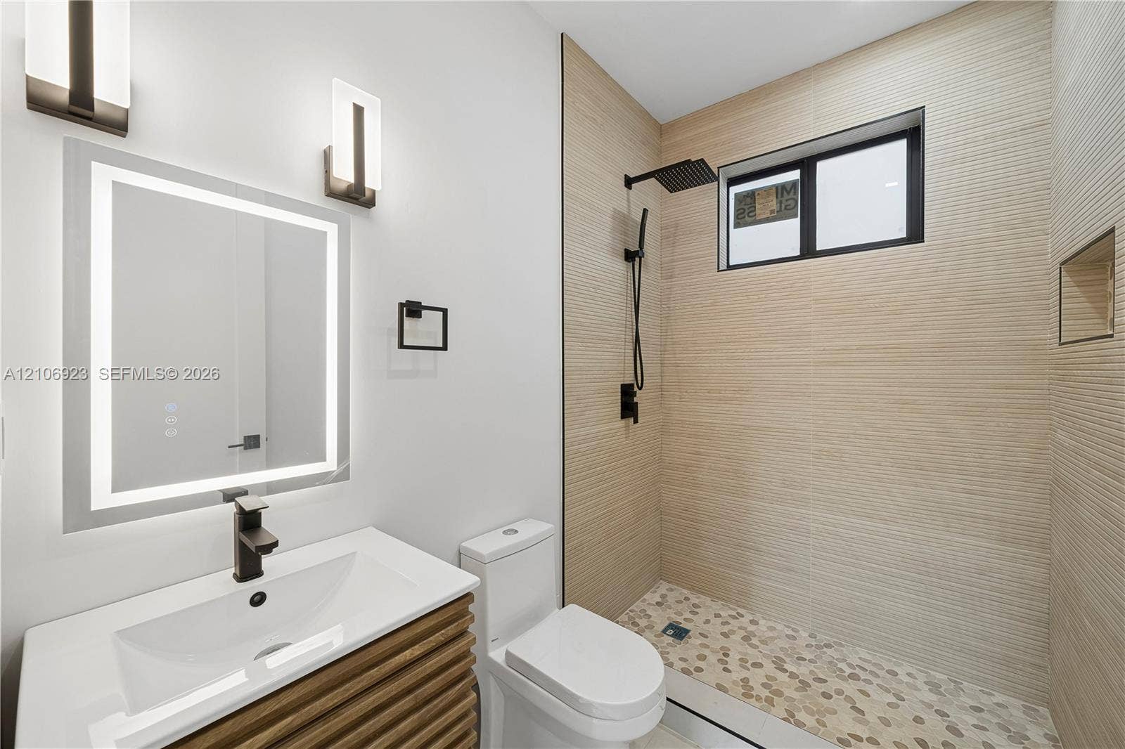 870 Superior Street, Opa Locka, FL 33054 Bathroom featuring a vanity with wood-finish cabinetry, a lighted mirror, matte black wall-mounted faucet, walk-in shower with textured tile walls, and a pebble-stone floor