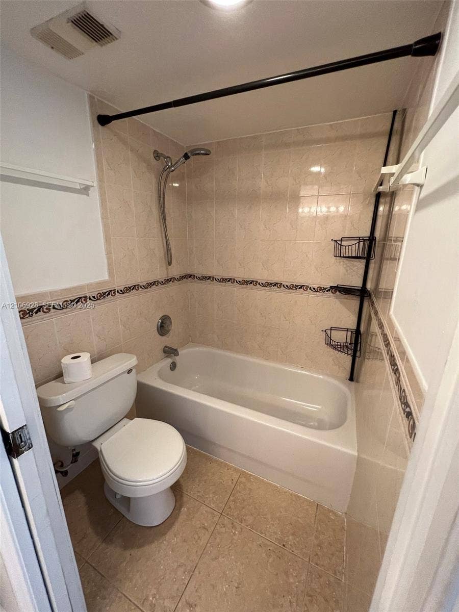 Bathroom featuring a tub-shower combination, neutral tiled walls with decorative accents, ceramic tile flooring, and a standard toilet
