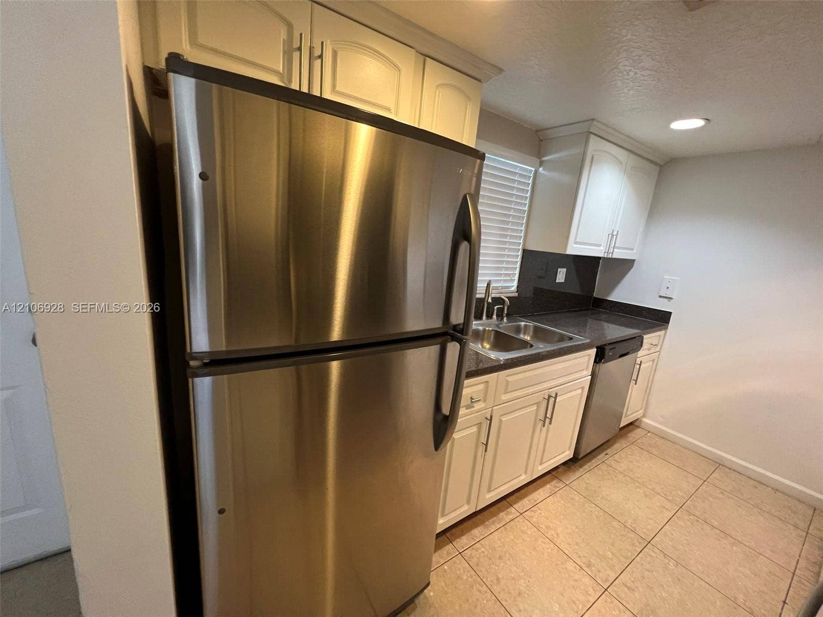 Kitchen featuring white shaker-style cabinetry, stainless steel refrigerator and dishwasher, dual-basin stainless steel sink, dark solid-surface countertops, and tiled flooring