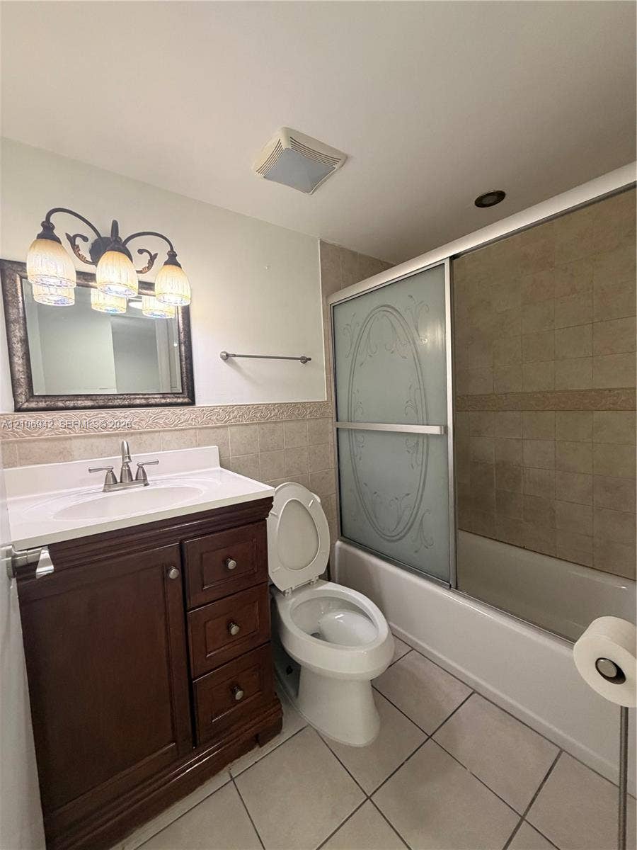 Bathroom featuring a dark wood-finish vanity with a white countertop, three-light decorative wall fixture, tile-surround bathtub with sliding glass doors, and light-colored tile flooring
