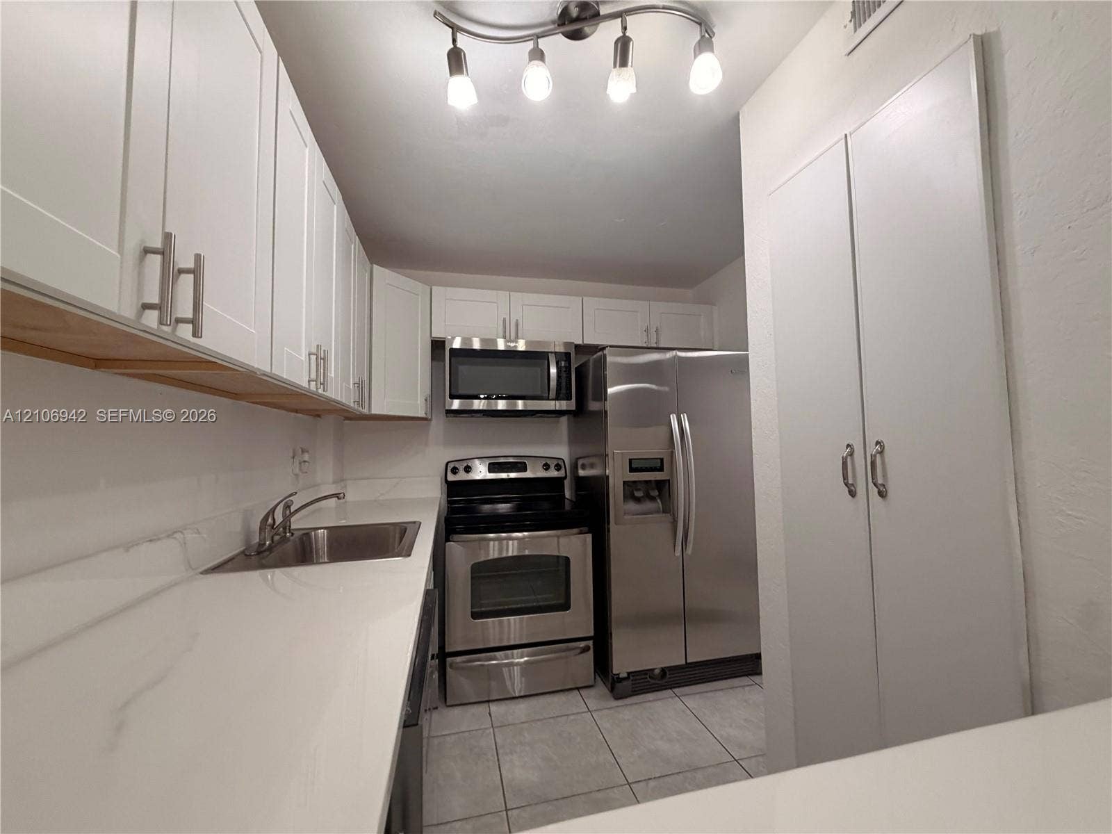 Kitchen featuring white shaker-style cabinetry, solid-surface countertops, stainless steel appliances, tile flooring, and multi-bulb track lighting