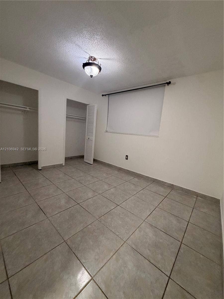 Neutral-toned ceramic tile flooring, two closets featuring white louvered doors, and a ceiling-mounted light fixture
