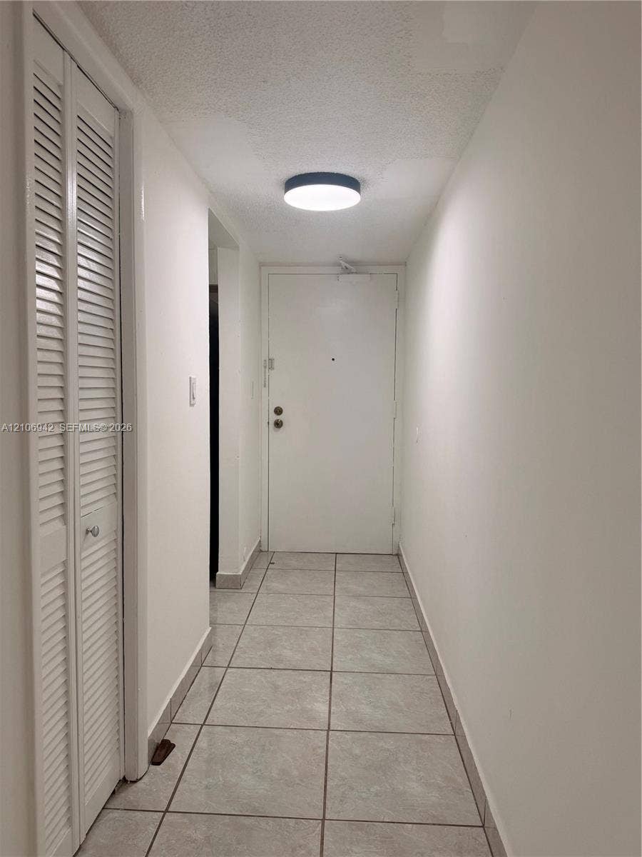 Neutral interior hallway featuring large-format ceramic tile flooring, floor-to-ceiling bi-fold closet doors with louvered panels, and a flush-mount circular LED light fixture