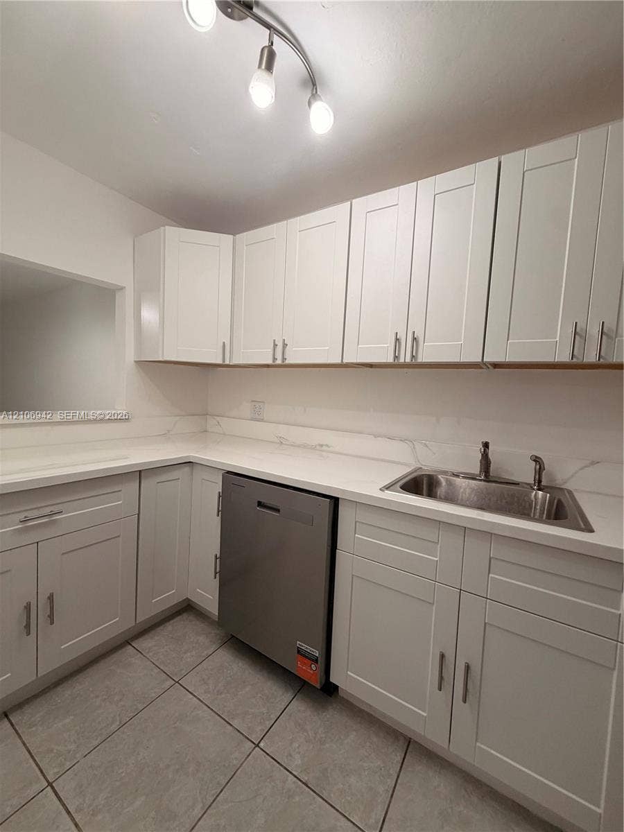 White shaker-style cabinetry, stone-finish countertops with integrated backsplash, stainless steel dishwasher, stainless steel sink with dual-handle faucet, and ceramic tile flooring