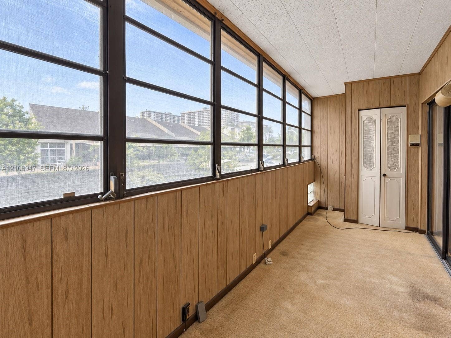 Enclosed space featuring vertical wood-finish wall paneling, a wall of multi-pane windows with metal framing, textured ceiling tiles, and dual-panel closet doors