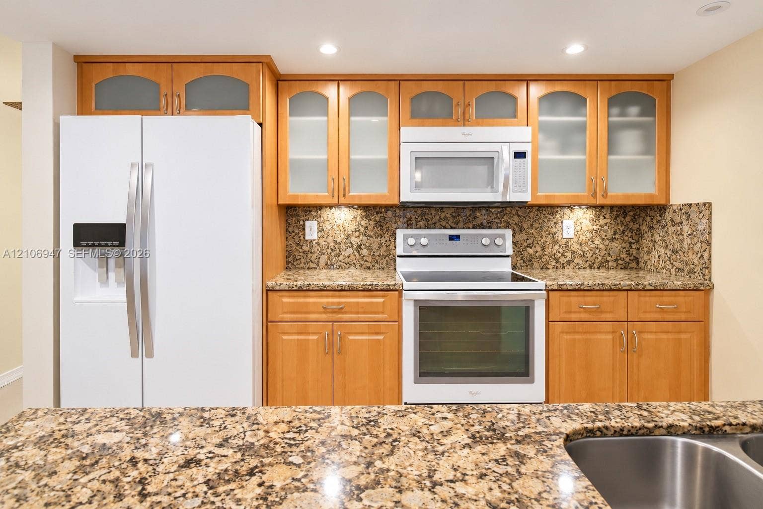 Kitchen featuring custom wood cabinetry with glass-panel upper doors, stone-finish countertops, stone-finish backsplash, and built-in white appliances