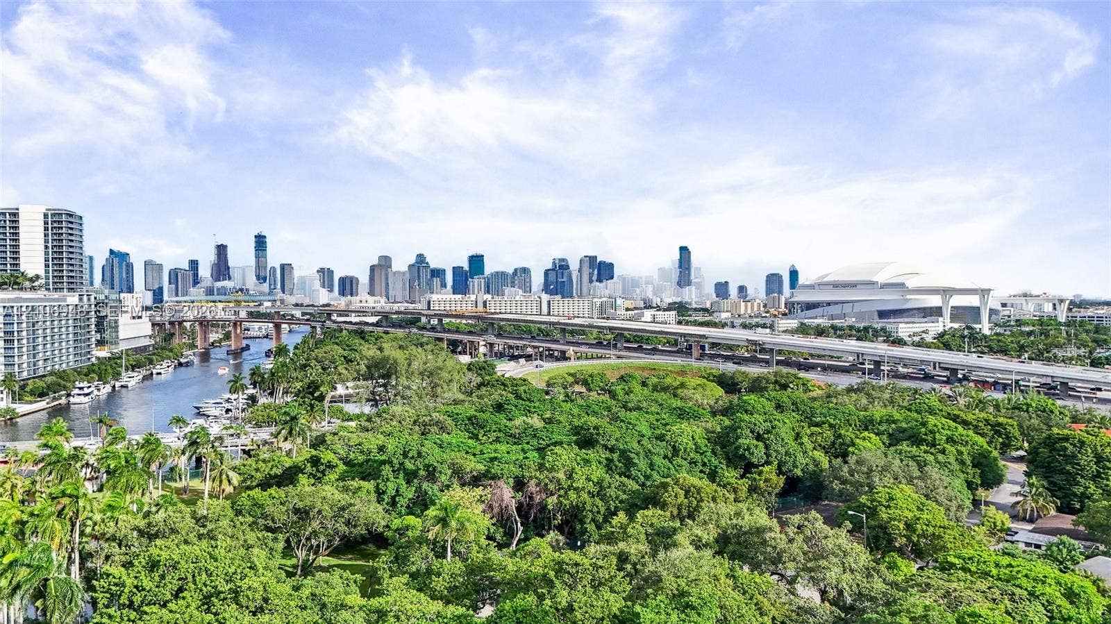 Expansive urban skyline, waterway with moored boats, dense green tree canopy, elevated roadways, and a prominent stadium structure