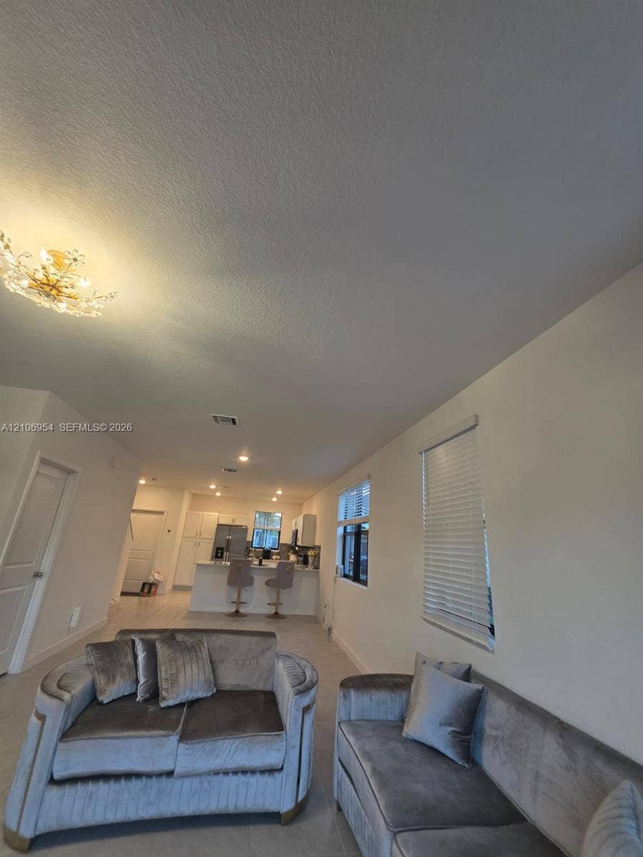 Open-concept interior featuring light-toned tile flooring, recessed lighting, ornate multi-bulb flush-mount chandelier, and windows fitted with horizontal blinds