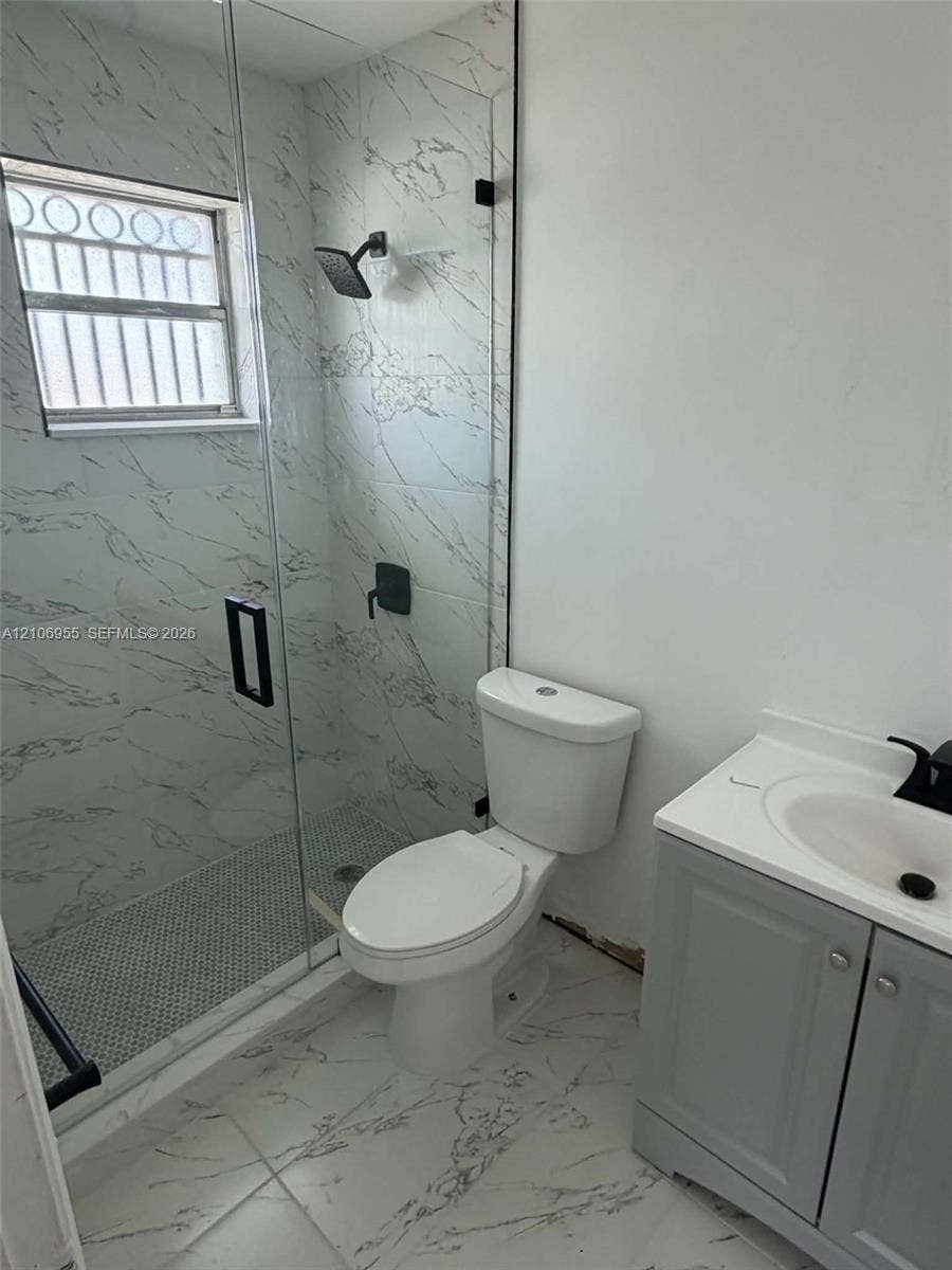 Walk-in shower featuring glass enclosure, wall-mounted matte black rainfall showerhead, and patterned tile surround, complemented by a grey vanity with white countertop and black fixtures