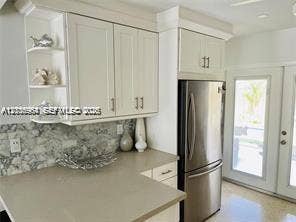 Kitchen featuring shaker-style cabinetry, open display shelving, neutral solid-surface countertops, stone-patterned backsplash, and stainless steel refrigerator