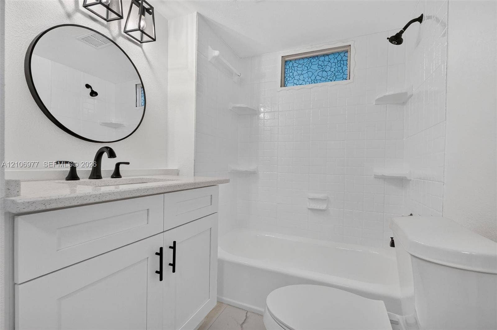 Bathroom featuring a white shaker-style vanity with stone-composite countertop, circular vanity mirror, matte black plumbing fixtures, and a tiled shower surround with integrated shelving
