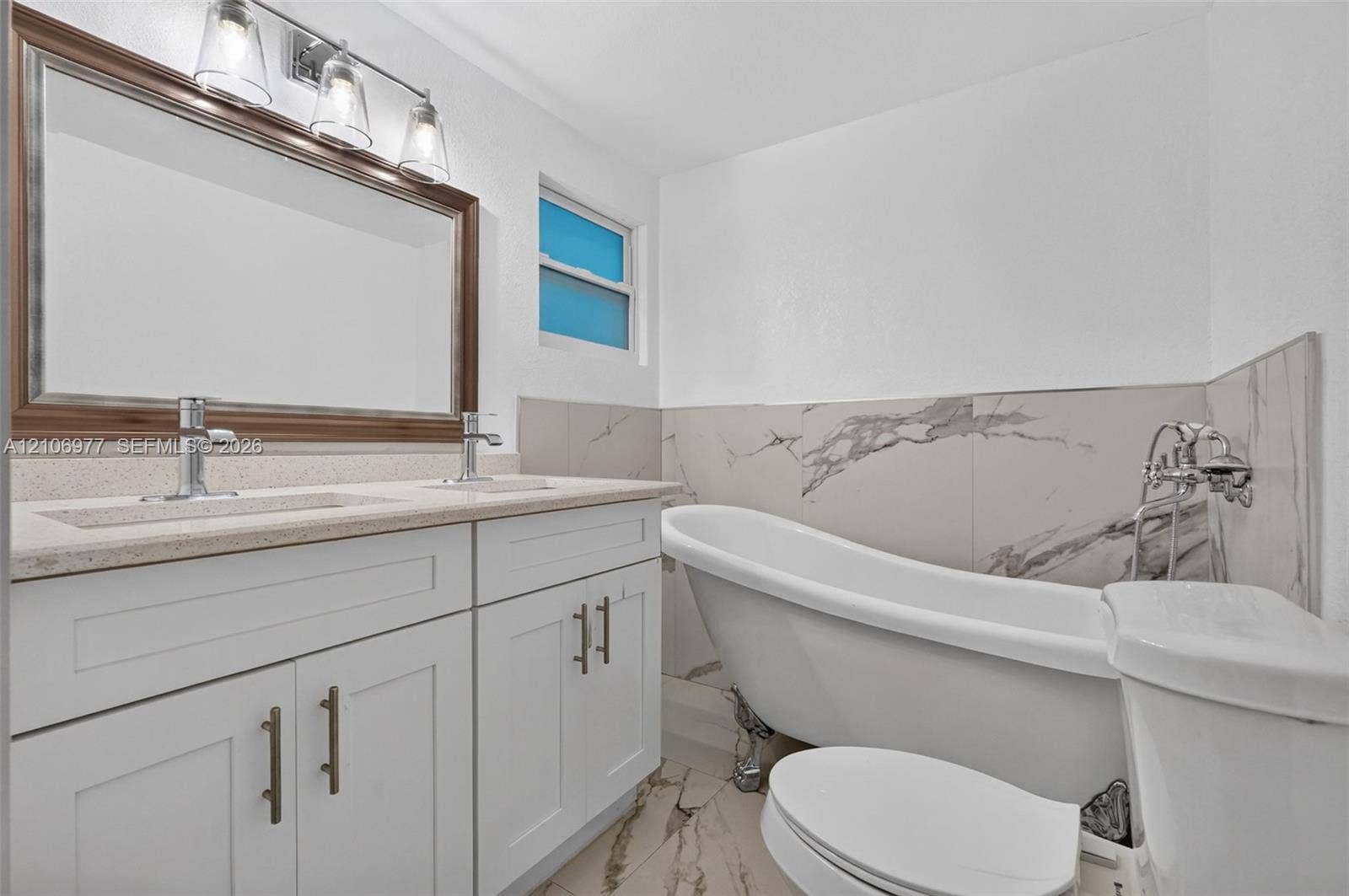 Dual-sink vanity with a stone countertop, freestanding clawfoot tub, large framed mirror with three-light fixture, and large-format tile wall accents