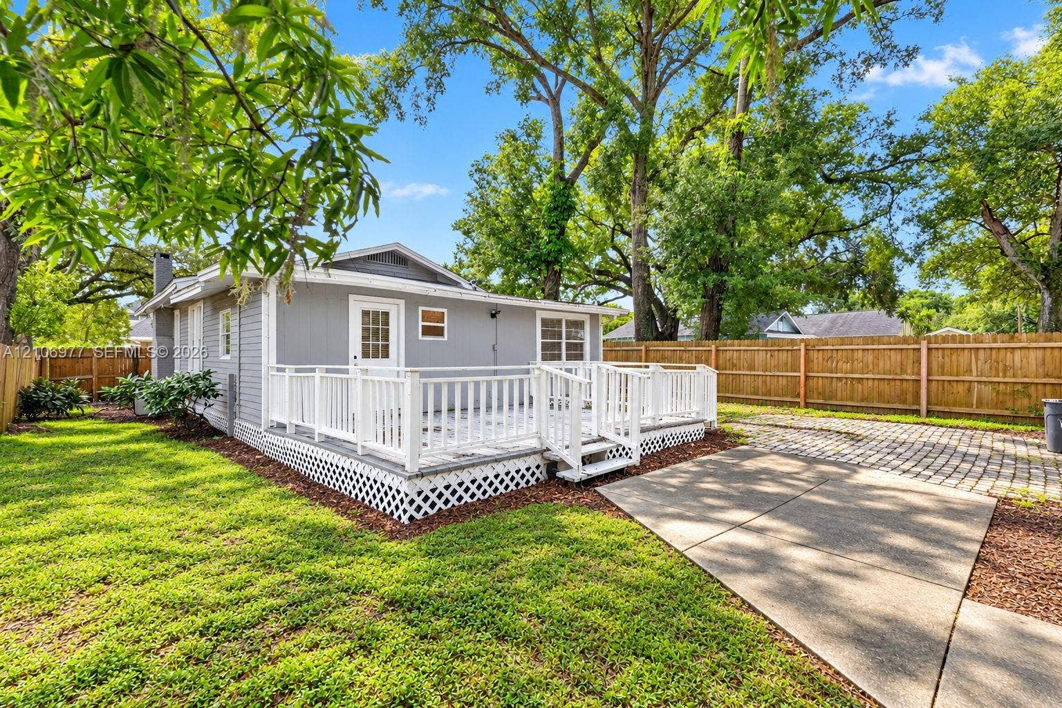 Exterior rear featuring a white wood-railing deck with lattice skirting, concrete patio, paver driveway, and wood-panel perimeter fencing set against mature trees