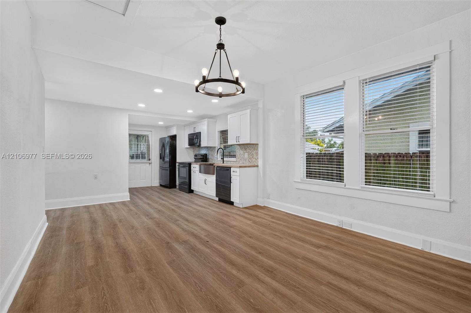 Open-concept living space featuring wood-finish flooring, white cabinetry, a farmhouse-style apron sink, matte black fixtures, and a statement chandelier