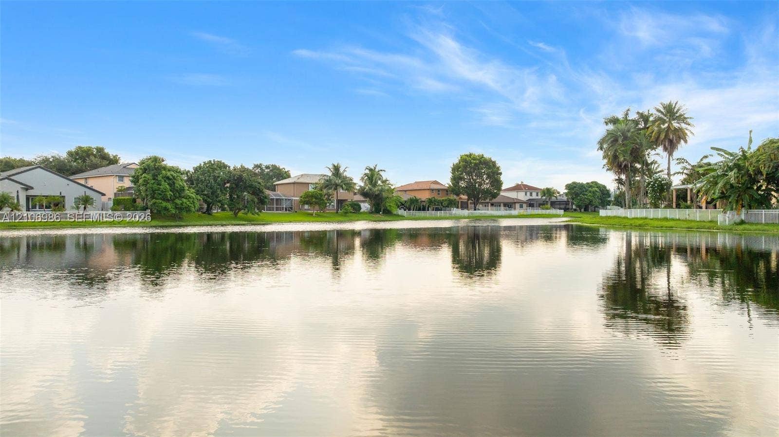 3801 SW 147th Avenue, Miramar, FL 33027 Waterfront setting featuring a calm pond, mature palm trees, perimeter white fencing, and expansive green space