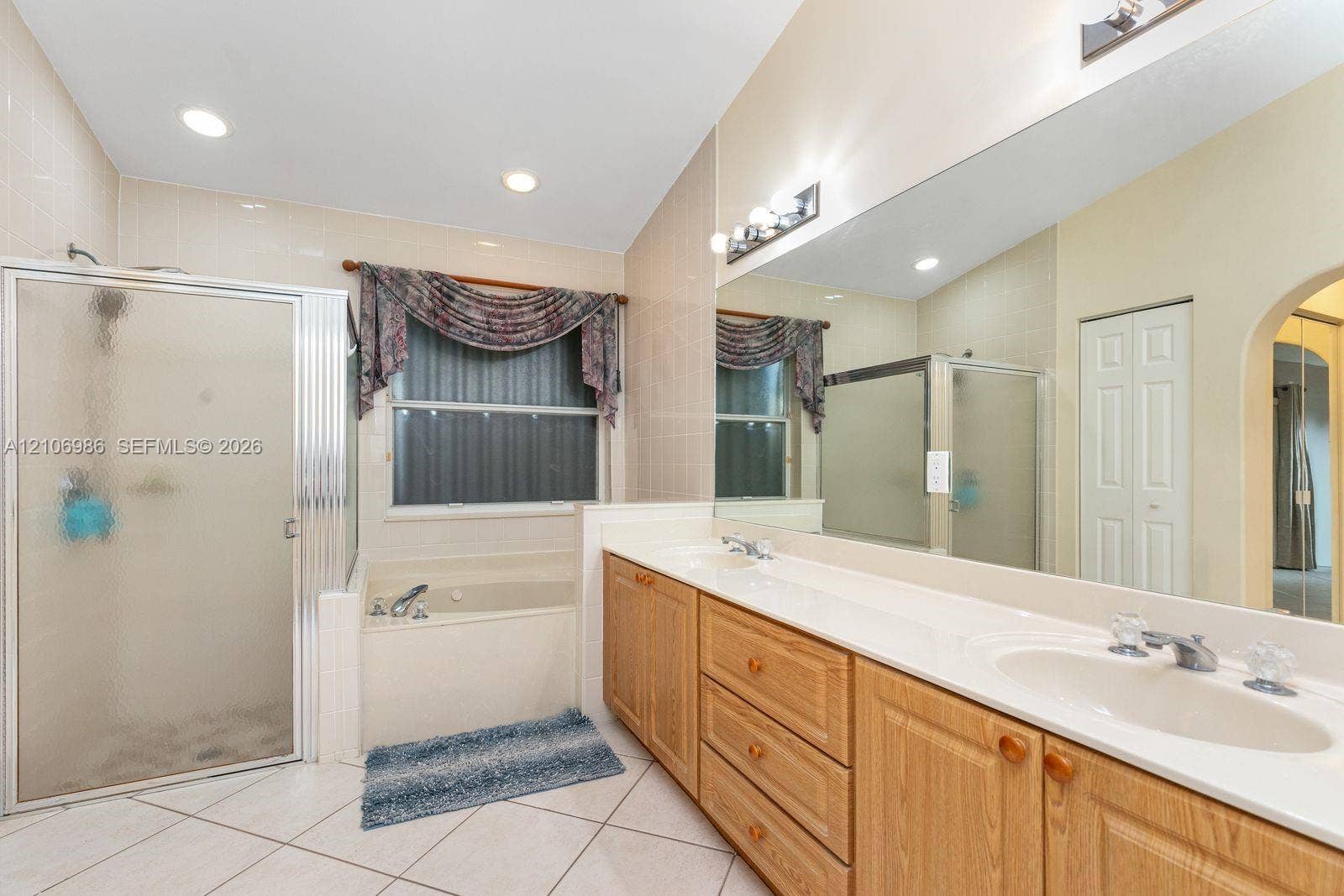 3801 SW 147th Avenue, Miramar, FL 33027 Bathroom featuring a dual-sink vanity with light wood cabinetry, a tiled-in soaking tub, a separate glass-enclosed shower, a full-width wall mirror, and tile flooring