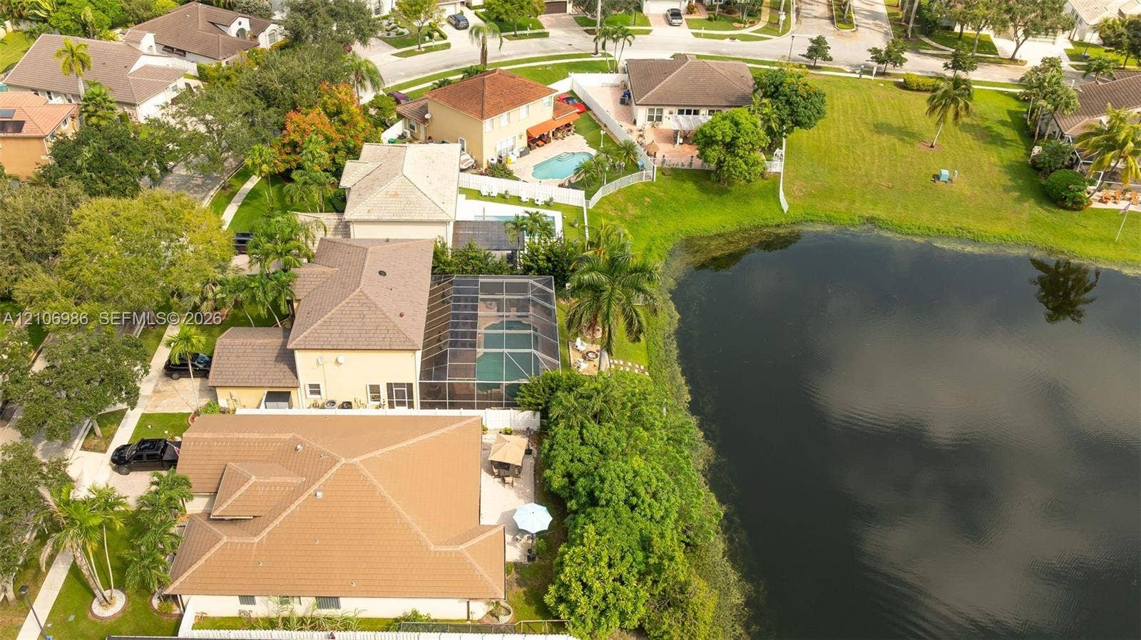 3801 SW 147th Avenue, Miramar, FL 33027 Aerial perspective featuring a waterfront property with a screened-in pool enclosure, concrete tile roofing, paved driveway, and mature landscaping