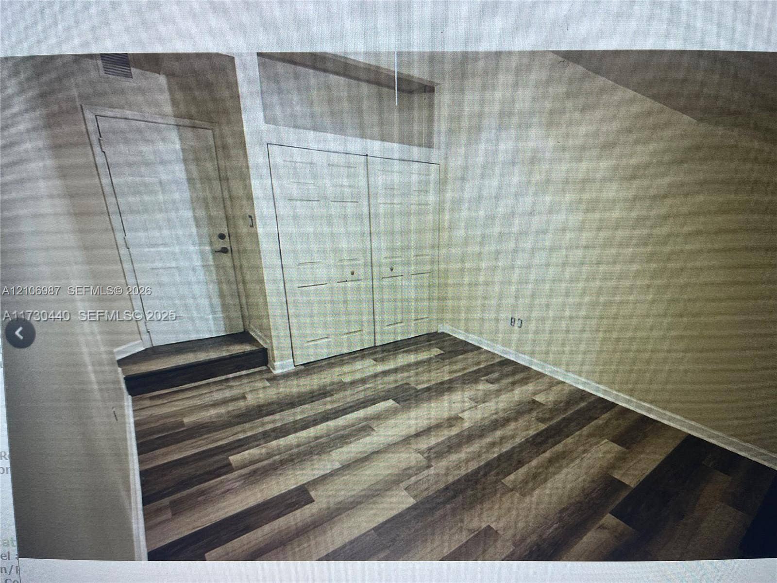 Room interior featuring wood-finish flooring, double-door closet with overhead built-in storage, and a single-panel door with a stepped entryway