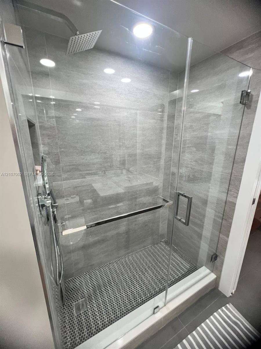 Walk-in shower featuring a frameless glass enclosure, floor-to-ceiling stone-finish tile surround, penny-round mosaic tile flooring, rain-style showerhead, and a handheld wand attachment