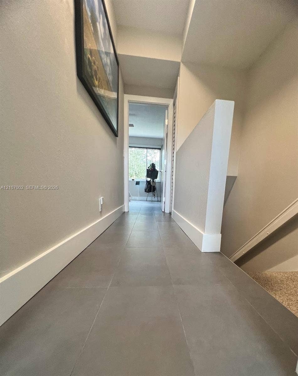 Hallway featuring large-format grey tile flooring, neutral wall paint, and partial stairwell wall