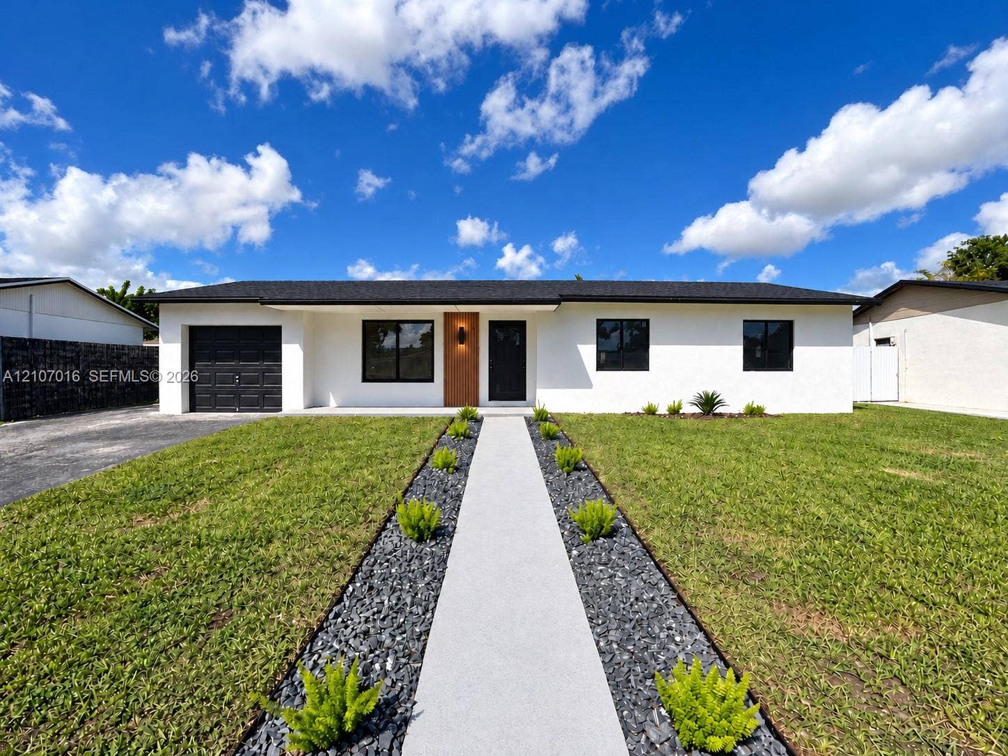 Single-story exterior featuring a stucco facade, dark-toned garage door, contemporary front door with wood-grain finish, asphalt shingle roof, and a paved walkway bordered by decorative stone and greenery